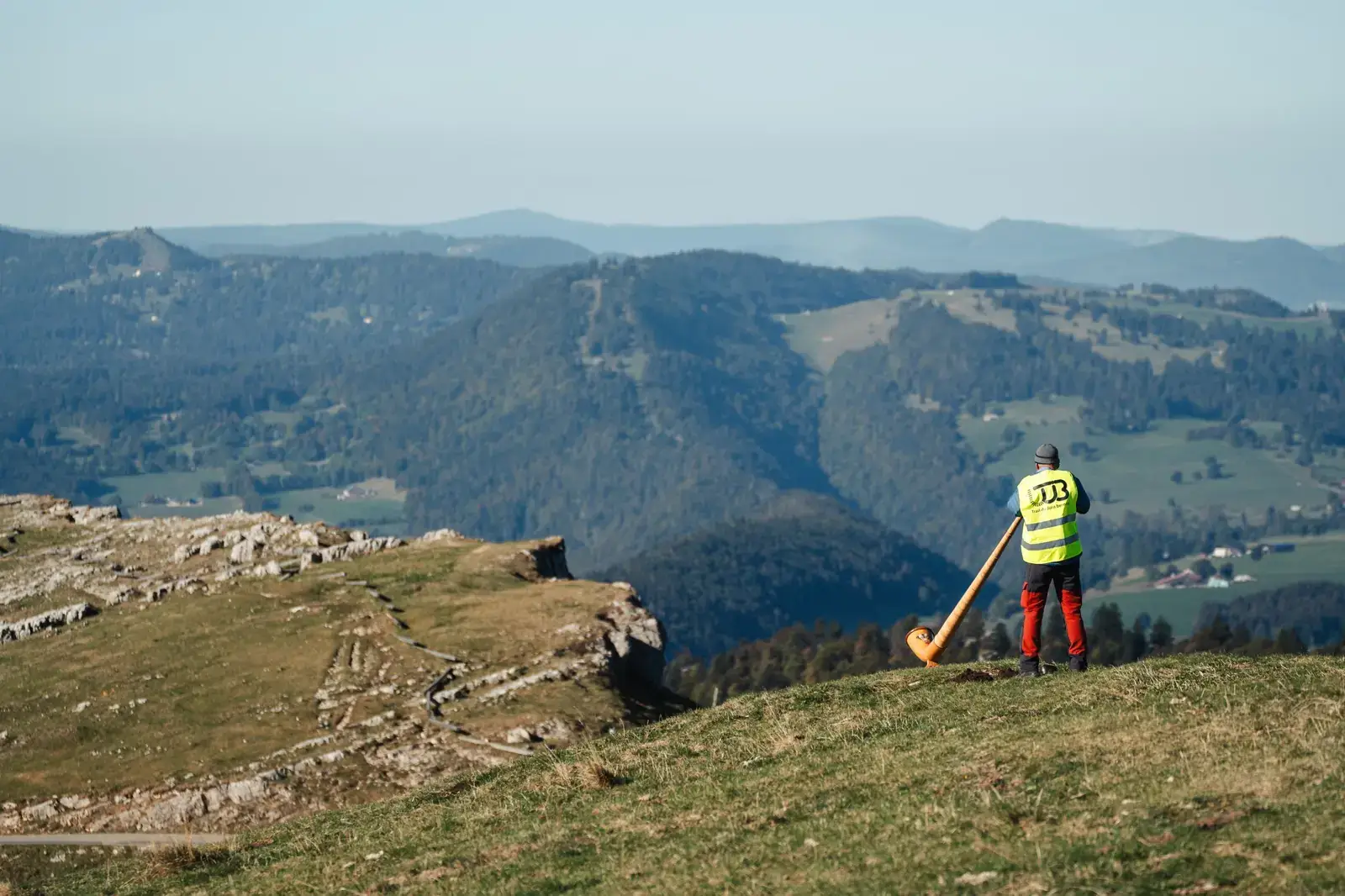 Ein Alphornbläser in gelber Weste auf einem Bergrücken, mit Blick auf die Täler des Juras